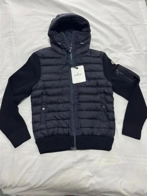 Moncler tricot cardigan - Snygg svart pufferjacka från Moncler med stickade ärmar och huva. Jackan har quiltad front, ribbade muddar och dragkedja framtill. Två fickor med tryckknappar och ikonisk Moncler-logga på ärmen. Perfekt för kalla vinterdagar.