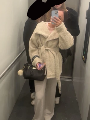 Beige kappa - Säljer en beige teddyjacka i mjukt, fluffigt material med bred krage och knytskärp i midjan. Jackan har en avslappnad passform och är perfekt för att hålla sig varm och snygg. Inga synliga loggor eller märken på jackan.