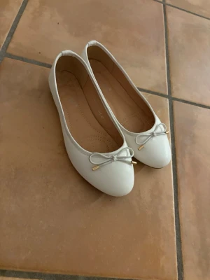 Vita ballerinaskor med rosett - Snygga vita ballerinaskor med rund tå och dekorativ rosett framtill. Skorna har en mjuk innersula och guldfärgade detaljer på rosetterna. Perfekta för vår och sommar kanske även student! , matchar enkelt till olika outfits. Använda en enda gång! Köpta på Raglady på väla centrum för 300kr!