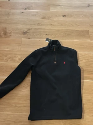 Svart half-zip tröja från Polo Ralph Lauren - Snygg svart långärmad tröja från Polo Ralph Lauren med half-zip och hög krage. Tröjan har det klassiska röda broderade logot på bröstet och är tillverkad i mjukt material som passar perfekt till en avslappnad stil. 