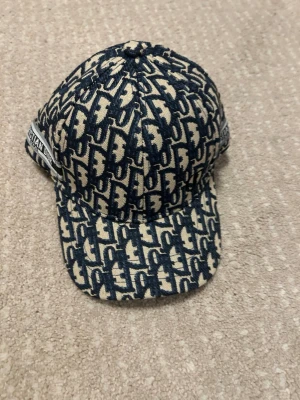 Christian Dior monogram keps blå/beige - Snygg keps från Christian Dior med klassiskt blått och beige monogrammönster över hela kepsen. Märket syns tydligt på sidan med en broderad etikett. Kepsen har böjd skärm och är tillverkad i ett tygmaterial.