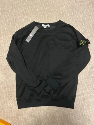 Svart sweatshirt från Stone Island - Snygg svart sweatshirt från Stone Island med den ikoniska loggan på vänster ärm. Tröjan har rund halsringning, ribbade muddar och är tillverkad i mjukt material som känns skönt mot huden. Perfekt för en clean och stilren look.