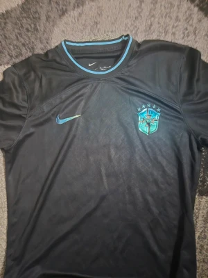 Svart Brasilien fotbollströja Nike - Snygg svart Brasilien fotbollströja från Nike med blå detaljer och CBF-emblem på bröstet. Tröjan har rund halsringning med blå kant och är tillverkad i ett lätt och sportigt material som passar perfekt till träning eller match.