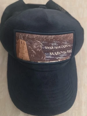 Svart Djurgården keps med patch - Svart keps med mesh baktill och justerbar snapback. Framtill sitter en stor patch med motiv av ett torn, en björn och texten 'Djurgår'n Hårskär'. Insidan har band med texten 'FORZA DJUUU'. Perfekt för dig som vill visa ditt Djurgården-supporterhjärta.