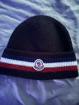 Svart Moncler mössa med ränder - Säljer en svart ribbstickad mössa från Moncler med bred uppvikt kant. Mössan har klassiska Moncler-ränder i blått, vitt och rött samt Moncler-logga framtill. Perfekt accessoar för kalla dagar och snygg till streetwear-stil.