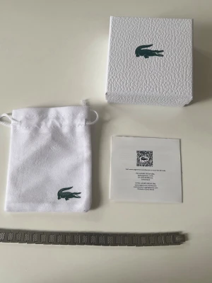 Silverfärgat armband från Lacoste - Snyggt armband i silverfärgad metall från Lacoste med fyrkantiga länkar och diskret Lacoste-logga. Kommer med original ask, tygpåse och certifikat. Perfekt accessoar för dig som gillar stilrena detaljer och ikoniska märken. Den är cirka 20 cm lång har en länkbortagare som kan anpassa till din önskade storlek.