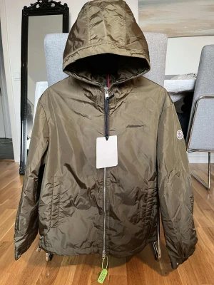 Olivgrön vindjacka från Moncler - Snygg olivgrön vindjacka från Moncler med huva och dragkedja framtill. Jackan har två sidofickor och Moncler-logga på ärmen. Tillverkad i ett lätt, glansigt material som står emot vind. Perfekt för dig som vill ha en stilren och funktionell jacka.