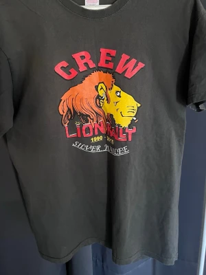 Svart t-shirt Lion Rally Crew - Svart t-shirt med stort tryck av ett lejon och texten 'CREW LION RALLY 1990-2015 Silver Jubilee' på framsidan. På baksidan står det 'Motorcycle Action Group' i gult. Rund hals och korta ärmar. Perfekt för dig som gillar unika tryck och bikerstil.