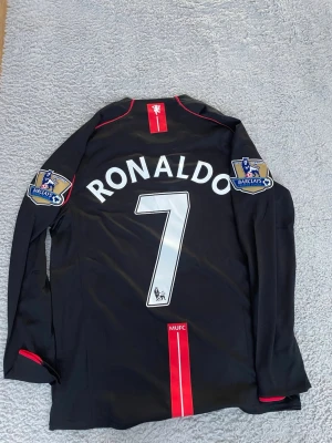 Manchester United Ronaldo tröja svart - Snygg svart långärmad Manchester United matchtröja från Nike med Ronaldo och nummer 7 på ryggen. Premier League-patchar på båda ärmarna, AIG som sponsor på bröstet och röda detaljer. Perfekt för fotbollsfans och samlare.
