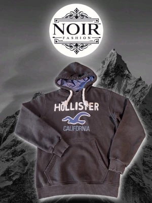 Svart Hollister hoodie med tryck - Svart hoodie från Hollister med stor vit och blå logga samt texten 'California' på bröstet. Utrustad med huva med blått innerfoder och vita dragsnören. Klassisk känguruficka framtill och ribbade muddar vid ärmslut och nederkant.