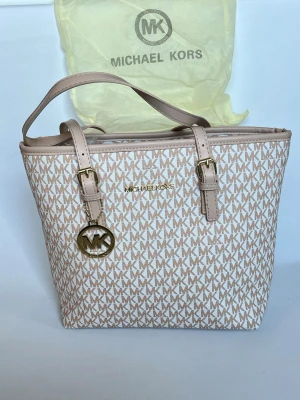 Beige Michael Kors handväska - Snygg handväska från Michael Kors i beige och ljusrosa toner med MK-logga över hela väskan. Väskan har två justerbara axelband, guldfärgade detaljer och en rund MK-berlock. Tillverkad i tåligt konstläder med klassisk, rymlig tote-modell.