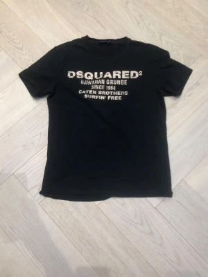 Svart Dsquared2 t-shirt med tryck - Säljer en svart t-shirt från Dsquared2 med vit text på bröstet. Klassisk rund hals och korta ärmar. Perfekt för dig som gillar streetwear och vill ha en enkel men snygg tee med coolt tryck framtill.
