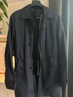 Mörkblå klassisk rock - Stilren mörkblå rock med knäppning framtill och två snedställda fickor. Rocken har en klassisk krage och justerbara ärmslut med knapp. Materialet ser ut att vara bomull eller en bomullsblandning, perfekt för kyligare dagar.