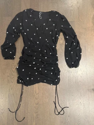 Svart prickig omlottklänning - Snygg svart kortklänning med vita prickar och omlottdesign. Klänningen har långa ärmar, snörning i sidorna och v-ringad hals. Perfekt för dig som gillar trendiga detaljer och en feminin look.