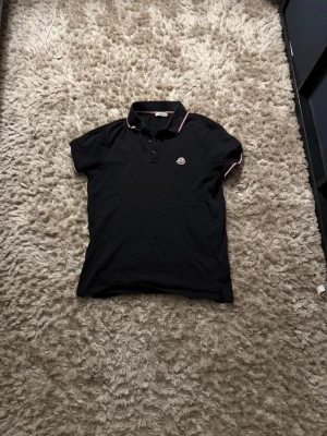 Svart Moncler pikétröja - Klassisk svart pikétröja från Moncler med korta ärmar och knappslå framtill. Tröjan har Moncler-logga på bröstet och diskreta vita och röda detaljer på krage och ärmslut. Perfekt till en avslappnad men snygg stil.