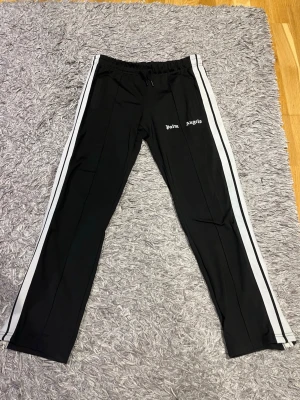 Palm Angels svarta mjukisbyxor - Palm Angels svarta mjukisbyxor med vita sidoränder och logga på vänster ben. Byxorna har resår i midjan och raka ben med dragkedjor vid bensluten. Tillverkade i ett mjukt och stretchigt material som ger en sportig vibe. Storlek M men passar även S