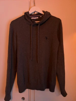 Mörkgrå hoodie från U.S. Polo Assn. - Mysig mörkgrå hoodie från U.S. Polo Assn. med broderad logga på bröstet. Tröjan har huva med dragsko och är tillverkad i mjukt stickat material. Perfekt för en chill och avslappnad stil.