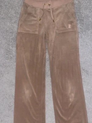 Beiga velourbyxor Juicy Couture - Säljer ett par beiga velourbyxor från Juicy Couture i storlek XS. Byxorna har bred resår i midjan med dragsko, två stora fickor fram och ett litet JC-emblem på ena fickan. Perfekta för chill dagar hemma eller när du vill vara extra comfy.