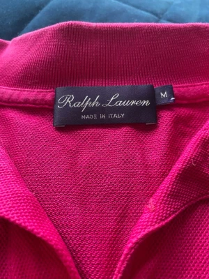 Rosa pikétröja från Ralph Lauren - Säljer en klassisk rosa pikétröja från Ralph Lauren, tillverkad i Italien. Tröjan har krage och den ikoniska broderade loggan med ryttare på bröstet. Materialet är piké i bomull som känns både bekvämt och stilrent.