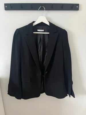 Svart kavaj från Mango - Stilren svart kavaj från Mango med klassisk passform och en knapp framtill. Kavajen har två fickor med lock och är helfodrad för en snygg siluett. Perfekt till en dressad look och enkel att matcha med olika outfits. Slits vid ärmarna 