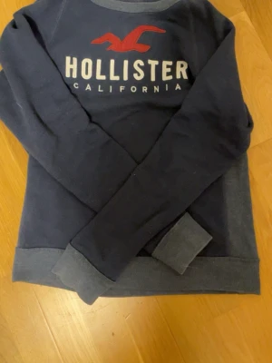 Mörkblå sweatshirt från Hollister - Mörkblå sweatshirt från Hollister med tryckt logga och text på bröstet. Tröjan har ribbade muddar vid ärmslut och nederkant samt rund halsringning. Perfekt för en avslappnad och stilren look.