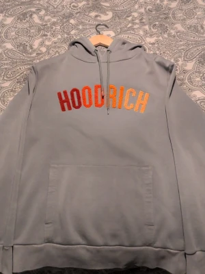 Grå HOODRICH hoodie med brodyr - Snygg grå hoodie från HOODRICH med broderad text i rött, orange och gult på bröstet. Klassisk känguruficka framtill och justerbar huva med dragsko. Perfekt för dig som gillar streetwear och vill ha en chill look.