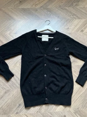 Svart kofta från Gant - Klassisk svart kofta från Gant med v-ringning och knäppning framtill. Broderad Gant-logga på bröstet. Långa ärmar och ribbade muddar. Perfekt att slänga över en t-shirt eller skjorta för en clean look.