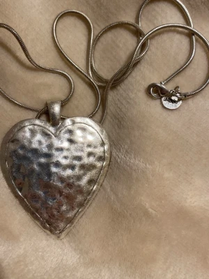 Silverfärgat hjärthalsband - Snyggt halsband med ett stort, hamrat hjärthänge i silverfärg. Kedjan är tunn och smidig med ett klassiskt lås. Perfekt accessoar för att lyfta vilken outfit som helst. 