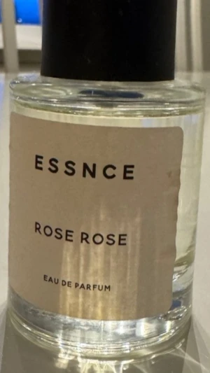 Essnce Rose Rose Eau de Parfum - En elegant parfymflaska från Essnce med doften Rose Rose. Flaskan är cylindrisk i genomskinligt glas med en beige etikett och svart kork. Doften är en Eau de Parfum och ger en fräsch och blommig känsla med tydliga rosnoter.