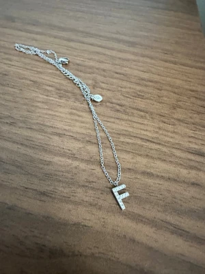Edblad Silverhalsband med F-bokstav - Snyggt halsband, från märket Edblad, i silverfärg med ett hängsmycke i form av bokstaven F. Bokstaven är dekorerad med små gnistrande stenar som ger ett elegant intryck. Kedjan är tunn och smidig, perfekt för dig som vill ha en personlig touch på din stil. Aldrig använt dock ingen originalförpackning, nypris 399:-
