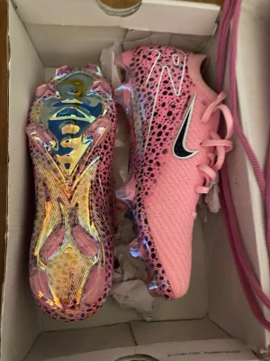 Nike rosa fotbollsskor med mönster - Säljer ett par riktigt snygga rosa Nike fotbollsskor med svart och vitt mönster samt holografisk sula. Skorna har snörning och är tillverkade i syntetmaterial för bästa prestanda på planen. Perfekta för dig som vill sticka ut på matchen! Dem är från en hemsida som säljer egendesignade Nike skor.