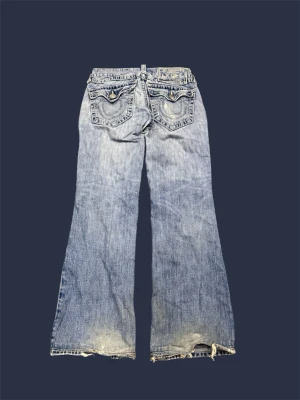 True Religion bootcut jeans - Säljer ett par ljusblå bootcut jeans från True Religion med slitna detaljer och stora bakfickor med dekorativa sömmar och knappar. Klassisk denimlook med logopatch bak i midjan och fransade benslut. Perfekta för dig som gillar vintage vibe. Storlek W31