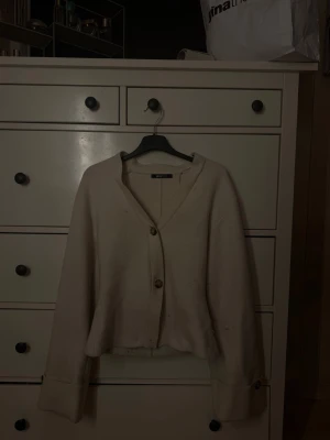 Beige kofta med stora knappar - Trendig beige kofta med v-ringning och stora bruna knappar framtill. Koftan har långa ärmar med breda muddar och en något boxig, croppad passform. Perfekt att slänga över en topp för en avslappnad look.