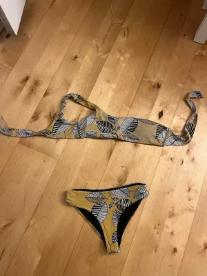 Gul bikini - Överdelen har långa knytband och en axel, medan underdelen är klassisk med samma mönster. Perfekt för stranden eller poolen! Överdelen är lite solblekt.