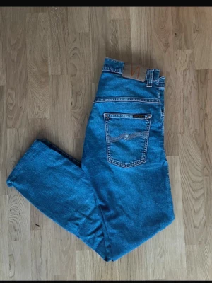Blå raka jeans från Nudie Jeans - Säljer ett par klassiska blå jeans från Nudie Jeans med raka ben och normal midja. Jeansen har fem fickor, tydliga sömmar och Nudie-loggan på bakfickan. Tillverkade i slitstark denim som ger en tidlös look. Perfekta till en avslappnad stil.