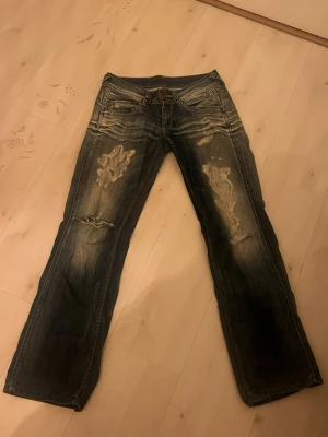 Blå slitna bootcut jeans (pepe jeans) - Säljer ett par blå bootcut jeans med slitningar och patchade detaljer på låren. Jeansen har klassisk femficksmodell, låg midja och tydliga tvättade effekter. Perfekta för en avslappnad och trendig look.