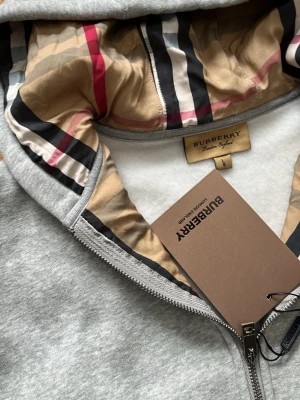 Grå zip hoodie från Burberry - Snygg grå zip hoodie från Burberry med klassiskt rutigt foder i huvan och diskret logga på bröstet. Hoodien har dragsko i huvan, fickor framtill och ribbade muddar. Perfekt för dig som vill ha en stilren och exklusiv look. Materialet känns mjukt och bekvämt.