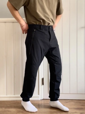 Svarta byxor från SOC - Säljer ett par svarta utomhus byxor från SOC i storlek L. Byxorna har dragkedjefickor på sidorna och en bekväm passform med resår vid bensluten. Materialet är mjukt och följsamt, perfekt för chill eller träning.