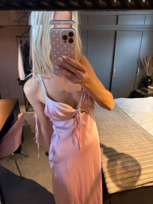 Ljusrosa satin långklänning  - Säljer en ljusrosa långklänning i satinliknande material med smala axelband och snygg snörning framtill. Klänningen har öppen rygg och v-ringning. Har en liten slitning vid ena sidan ryggen, men inget man tänker på. Prislapp kvar.