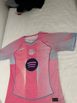 Barça x Nike rosa matchtröja - Snygg FC Barcelona matchtröja i rosa och ljusblått från Nike. Tröjan har korta ärmar, V-ringning med turkos kant och klubbmärke samt Nike-logga på bröstet. Tillverkad i lätt, ventilerande material perfekt för träning eller match.