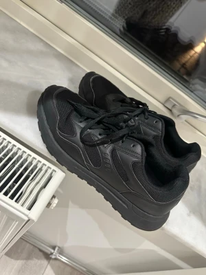  svarta sneakers med meshdetaljer - Säljer ett par helsvarta sneakers med snygga meshdetaljer och robust sula. Skorna har klassisk snörning och en sportig look som passar till det mesta. Perfekta för dig som gillar stilrena och bekväma skor till vardags.