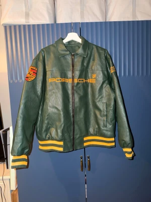 Grön Porsche bomberjacka  - Säljer en grön bomberjacka i skinn från Porsche med gula detaljer. Jackan har broderade Porsche-loggor fram och bak, stickade muddar i gult och grönt och dragkedja framtill. Perfekt statement-plagg för bilentusiasten. OBS DETTA ÄR INTE EN ALD JACKA!