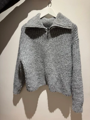 Grå stickad ziptröja med krage - Mysig grå stickad tröja med grov struktur och dragkedja framtill. Tröjan har en bred krage som kan bäras både uppvikt och nedvikt samt långa ärmar med ribbade muddar. Perfekt för lager på lager och chill stil.