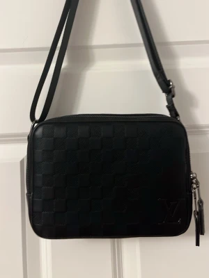 Svart axelväska Louis Vuitton - Snygg svart axelväska från Louis Vuitton med klassiskt rutmönster och präglad LV-logga på framsidan. Väskan har justerbar axelrem och detaljer i silverfärgad metall. Tillverkad i läder med stilren och modern design.