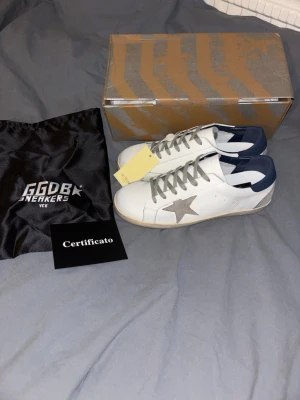 GGDB Superstar vita sneakers - Säljer ett par GGDB Superstar sneakers i vitt läder med grå stjärna på sidan, grå skosnören och mörkblå häl. Klassisk låg modell med slitstark gummisula och detaljerad branding på plös och häl. Kommer med originalkartong, dustbag och certifikat.