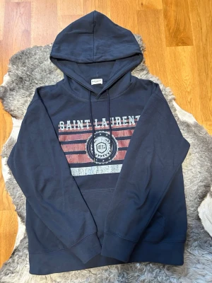 Mörkblå hoodie från Saint Laurent - Snygg mörkblå hoodie från Saint Laurent med tryck framtill i vitt och rött samt texten 'SAINT LAURENT'. Klassisk huva med dragsko och känguruficka. Tillverkad i mjukt bomullsmaterial som känns skönt mot huden. Perfekt för en avslappnad och trendig look.