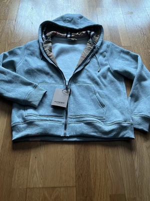 Grå zip hoodie från Burberry - Snygg ljusblå hoodie från Burberry med dragkedja och klassiskt rutigt foder i huvan. Hoodien har känguruficka framtill, ribbade muddar och en avslappnad passform. Perfekt för dig som vill ha en stilren och exklusiv look.
