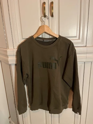 Olivgrön sweatshirt från Puma - Olivgrön sweatshirt från Puma med klassisk logga framtill i mörkare ton. Tröjan har rund halsringning, långa ärmar och ribbade muddar vid ärmslut och nederkant. 
