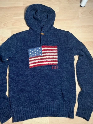 Blå Polo Ralph Lauren Hoodie - Säljer nu en blå stickad hoodie från Polo Ralph Lauren med amerikansk flagga på bröstet och RL67-brodyr.   Tröjan är i utmärkt skick och har inga defekter.  Är i ungdomsstorlek i Large, vilket motsvarar Xs/S i vuxen storlek.           Längd: Armhåla till armhåla - 47 cm.  Läng på hoodien - 57 cm.                                                                            Nypris ca 2000 kr - mitt pris 849 kr.  Priset är inte hugget i sten. Skriv gärna om ni har några mer funderingar🙂/Filip