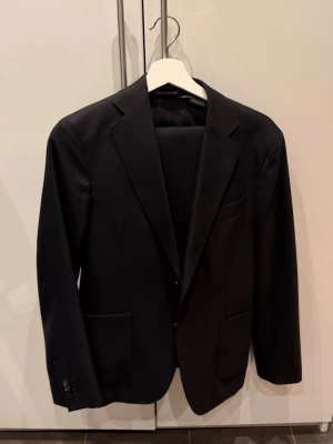 Svart Kostym från Oscar Jacobsson - Stilren svart kavaj med två knappar framtill och smala slag. Kavajen har tre fickor, varav en bröstficka, och knappar vid ärmsluten. Perfekt för en dressad look och enkel att matcha med skjorta eller t-shirt.